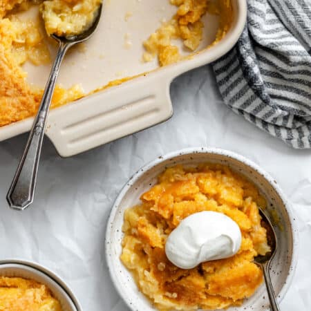 A bowl of pineapple dump cake topped with whipped cream next to the cake baking pan.