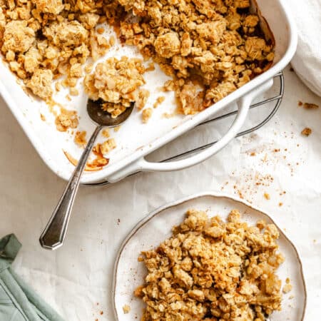 A plate of apple crisp next to the baking dish of crisp with a spoon in it.