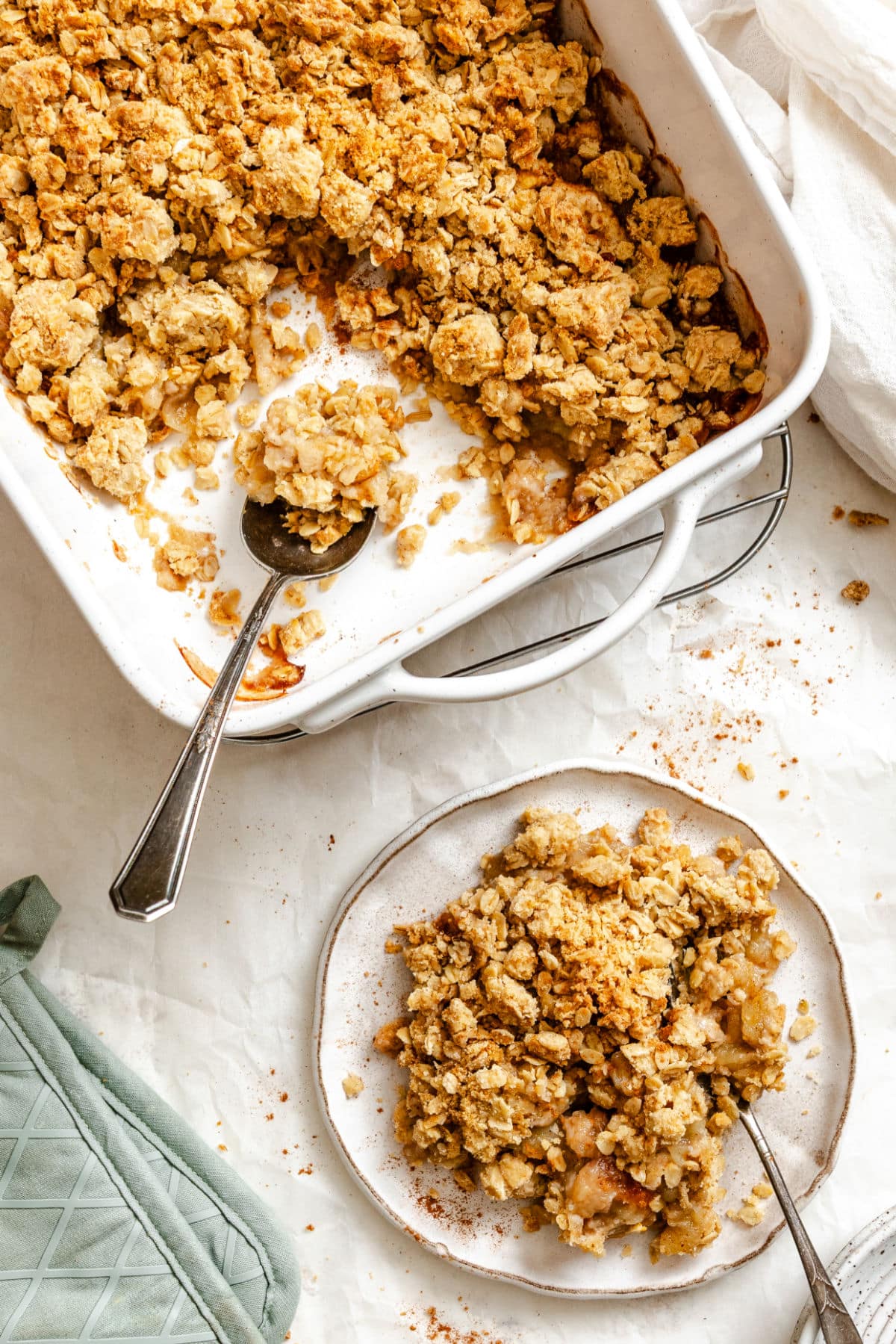 A plate of apple crisp next to the baking dish of crisp with a spoon in it. 