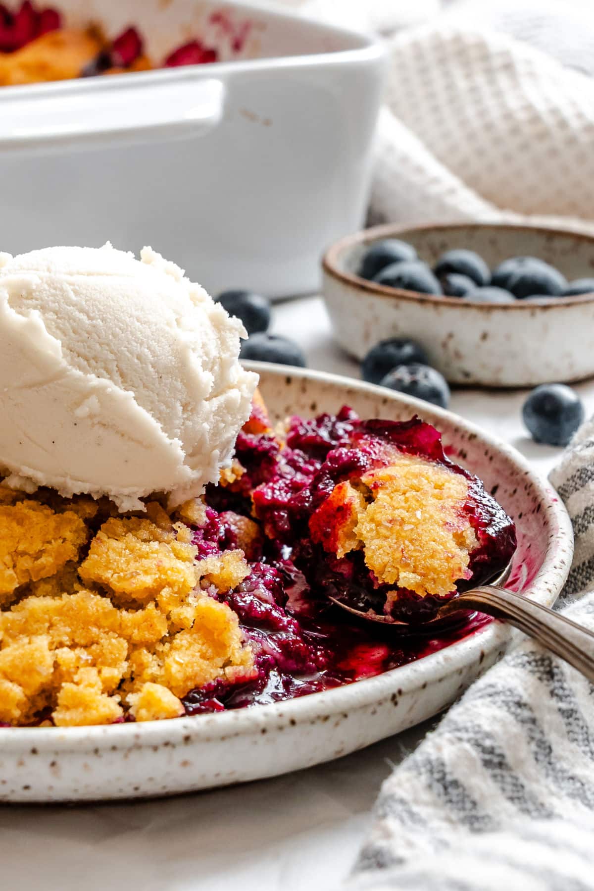 A bowl of blueberry cobbler topped with vanilla ice cream with a spoon in it.