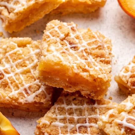 Two peach crumble bars leaning against each other surrounded by bars and fresh peaches.