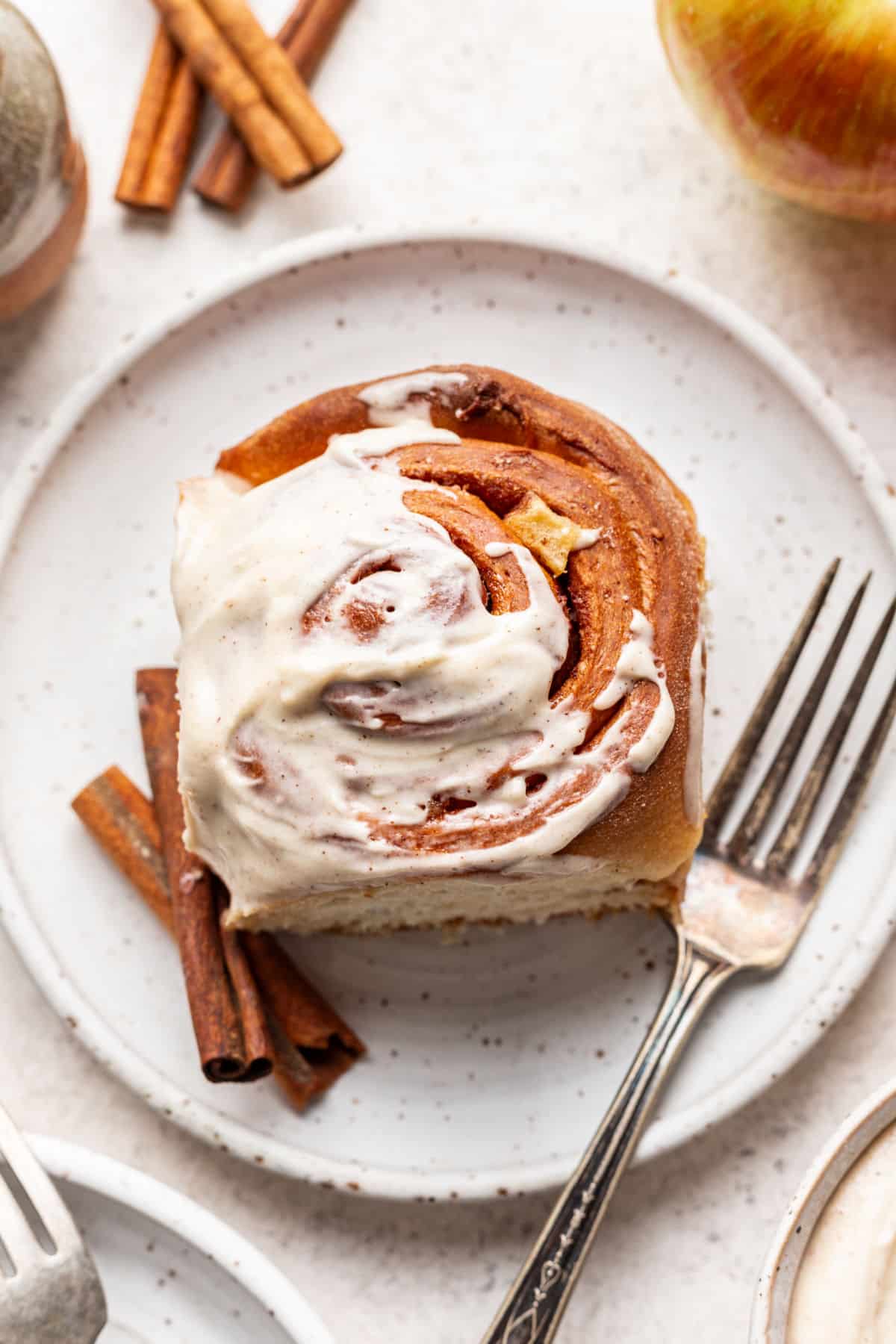 A partially frosted apple cinnamon roll on a plate next to a fork and cinnamon sticks.