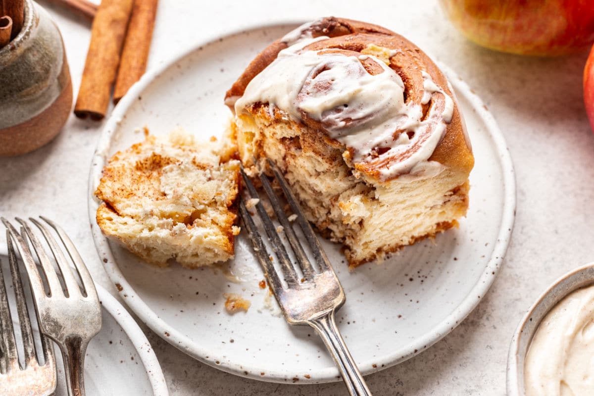 A fork taking a bite of an apple cinnamon roll on a plate.
