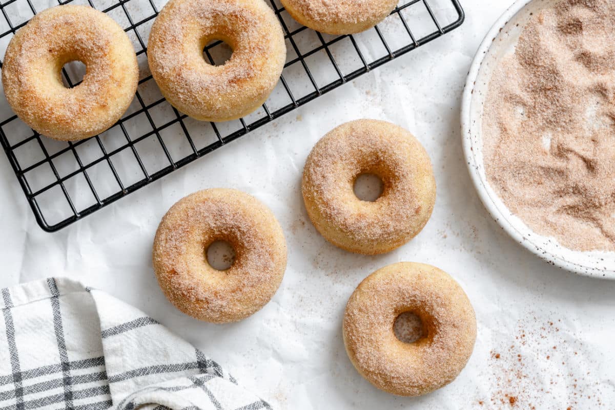 Cinnamon Sugar Baked Donuts Baked cinnamon sugar donuts on white parchment paper next to a dish of cinnamon sugar.