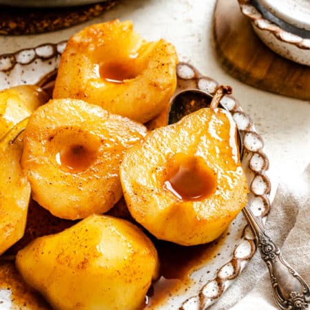 Cider poached pears on a speckled plate next to a silver spoon.