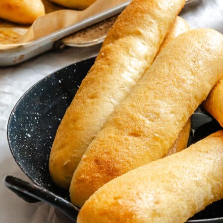 Three garlic breadsticks on a black platter next to a pan of breadsticks.