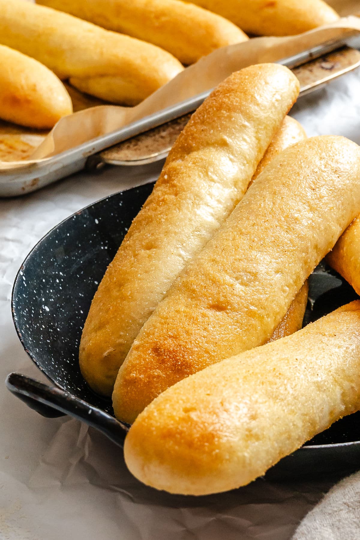 Three garlic breadsticks on a black platter next to a pan of breadsticks.