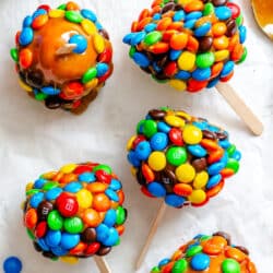 M&M caramel apples on a piece of white parchment paper next to a dish of caramel.