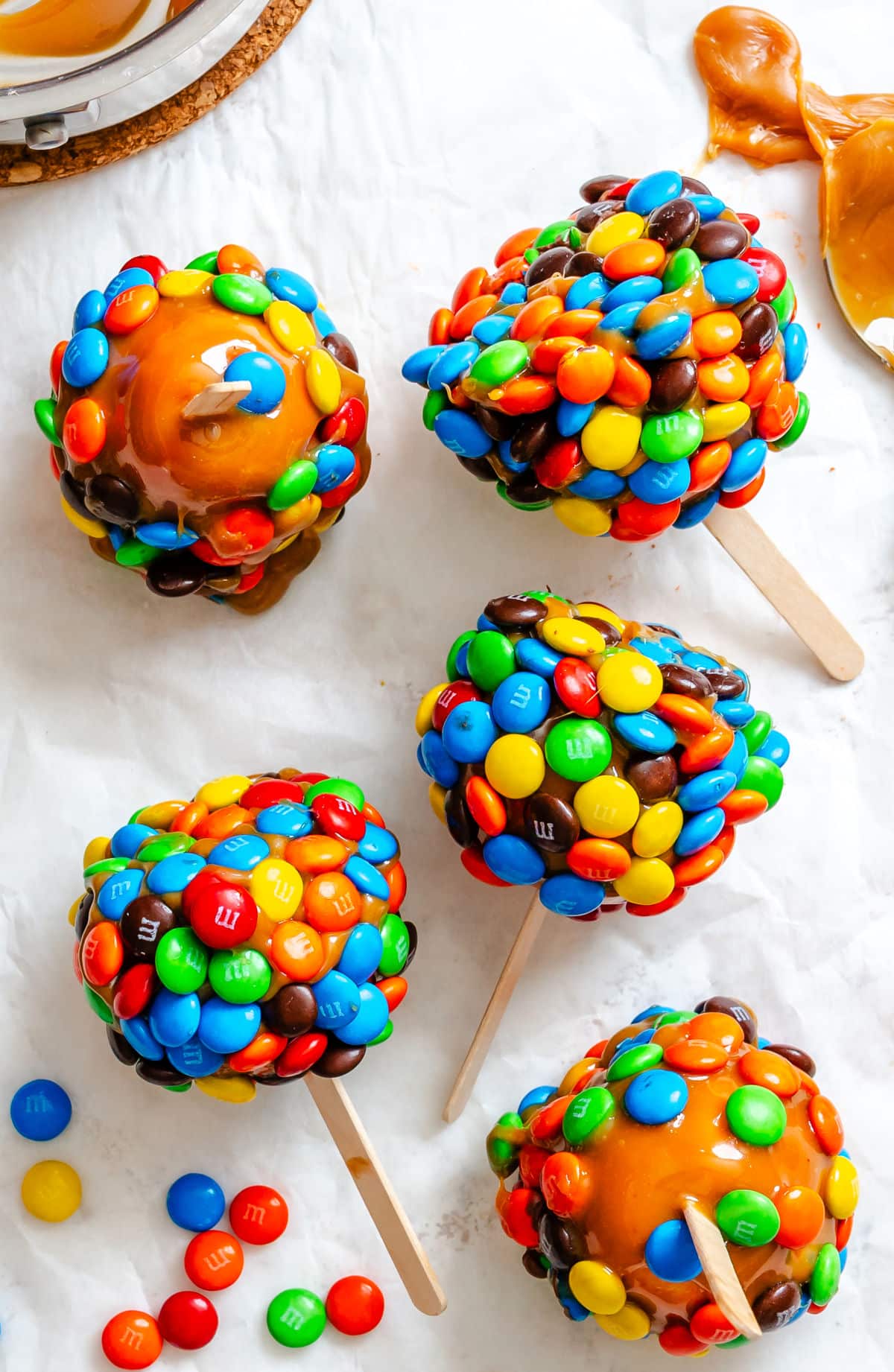 M&M caramel apples on a piece of white parchment paper next to a dish of caramel.