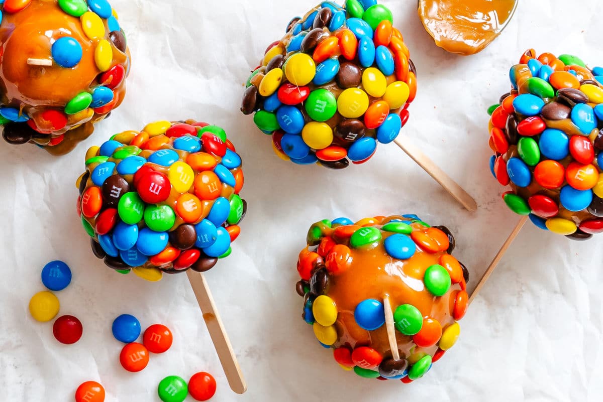 Four M&M caramel apples next to M&MS on white parchment paper.