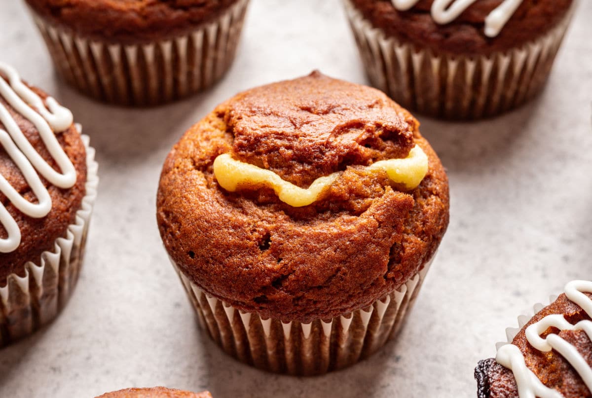 An uniced pumpkin cream cheese muffin next to iced muffins.
