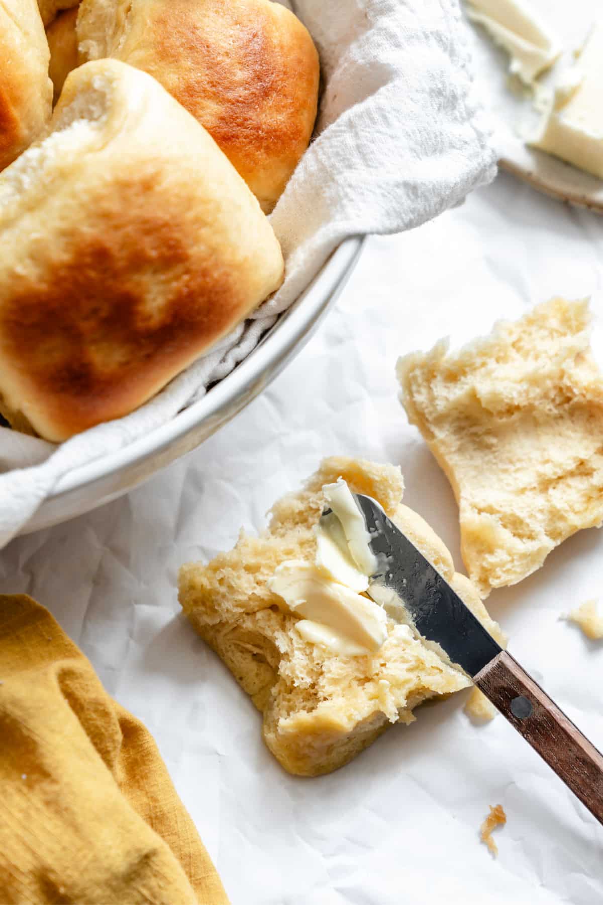 A knife spreading butter onto a 1 hour Hawaiian roll that's cut in half.