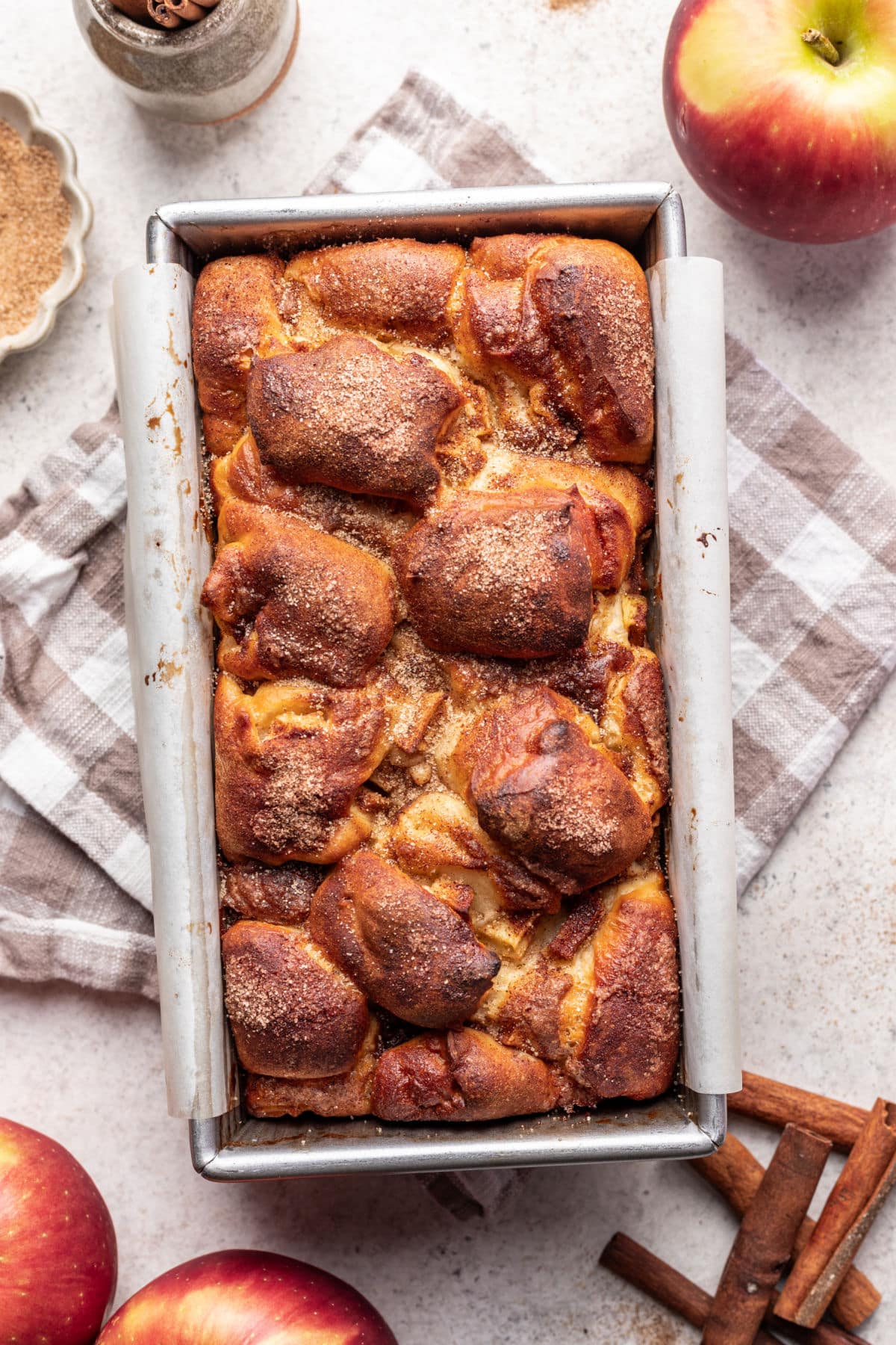 A baked loaf of apple cinnamon bread in the pan next to a tan checkered kitchen towel and apples. 