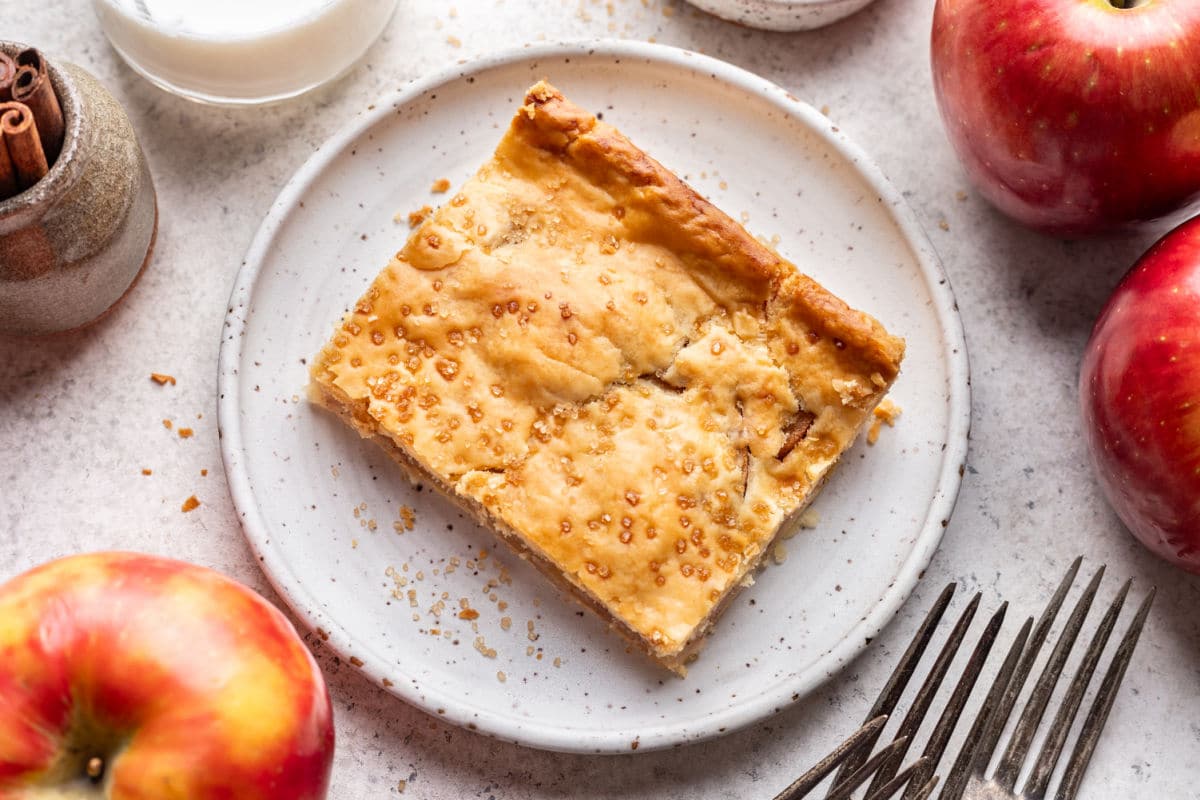 A slice of apple slab pie on a plate surrounded by apples. 