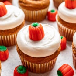 Pumpkin cupcake with cream cheese frosting topped with a candy pumpkin next to more candy pumpkins.