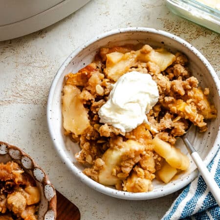 A dish of slow cooker apple crisp topped with whipped cream next to a kitchen towel.