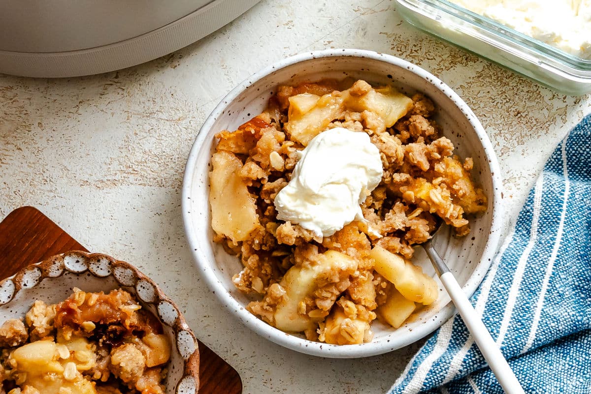 A dish of slow cooker apple crisp topped with whipped cream next to a kitchen towel.