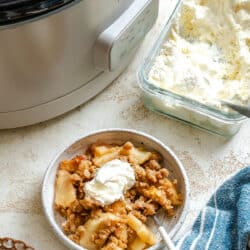 A dish of slow cooker apple crisp next to a dish of homemade whipped cream.
