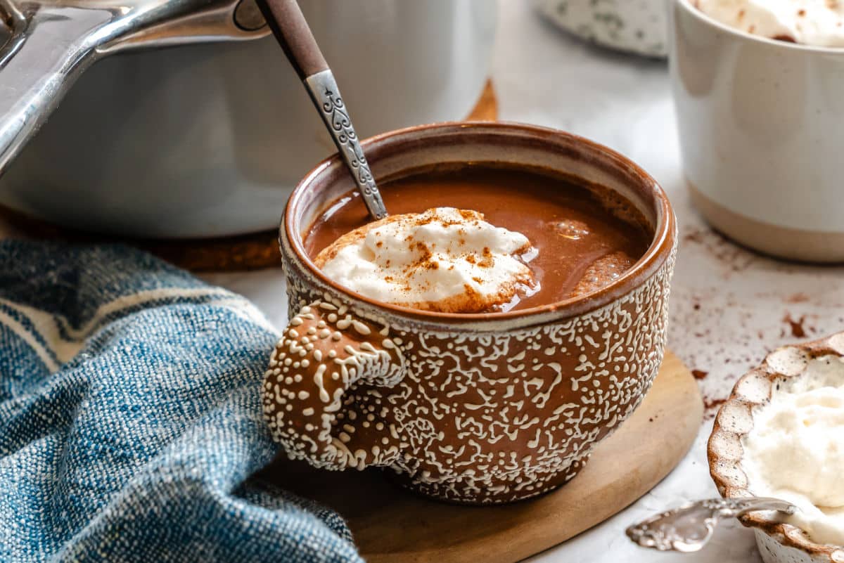 A mug of gingerbread hot chocolate with a spoon in it next to the saucepan.