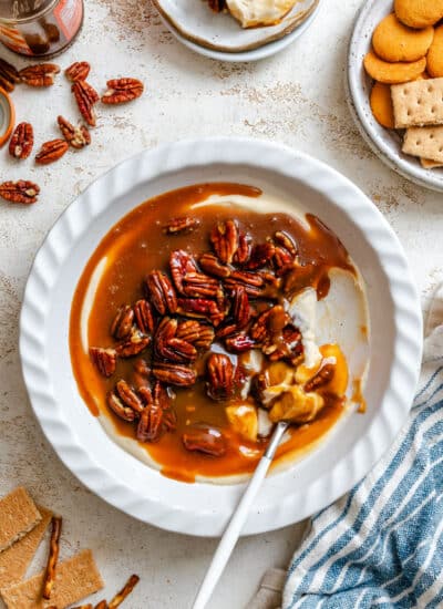 A dish of pecan pie dip with a spoon in it next to scattered pecans.