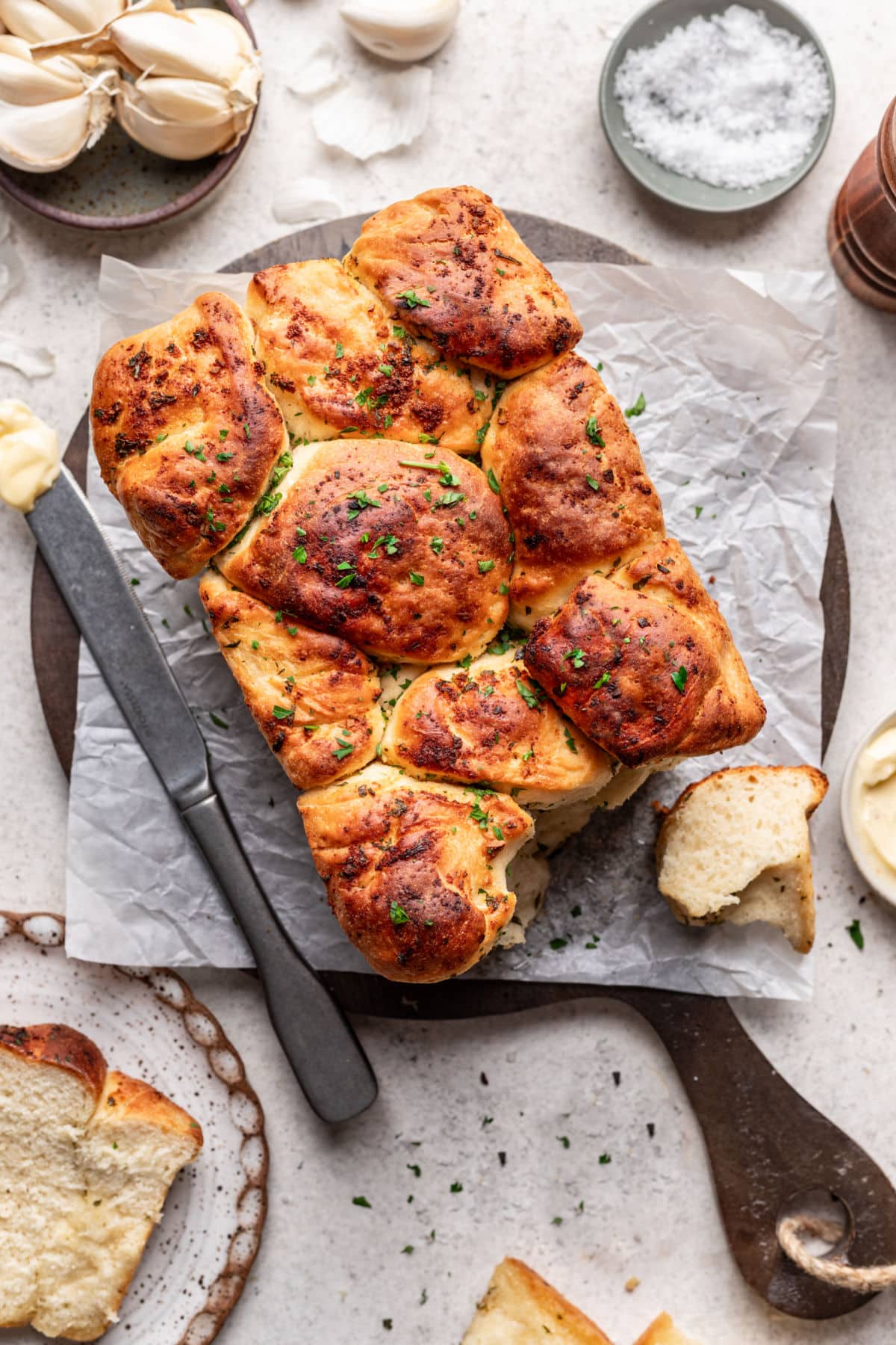 A loaf of garlic pull apart bread on a cutting board next to dishes of butter and salt.