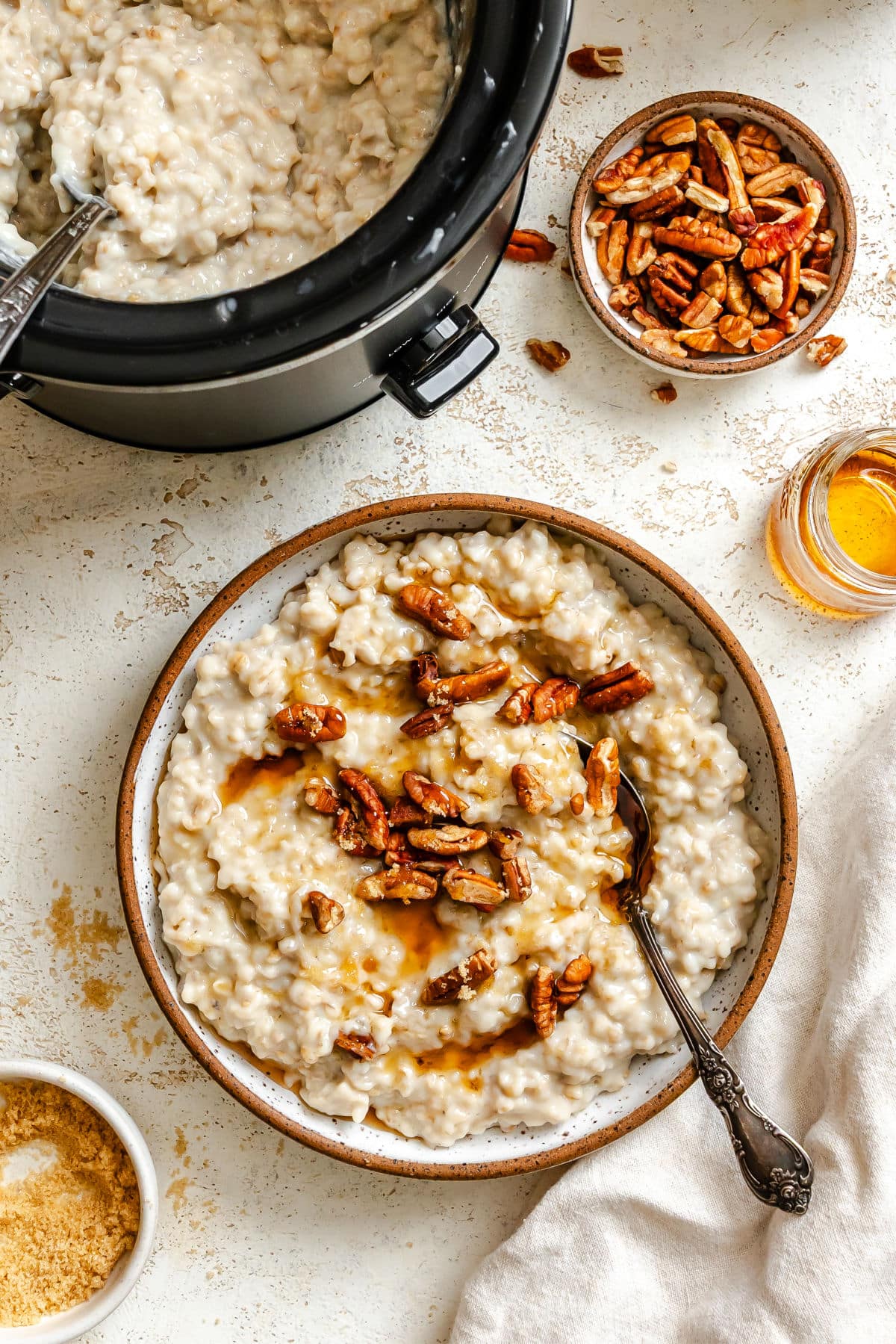 A bowl of slow cooker pecan pie oatmeal next to oatmeal in the crock pot. 