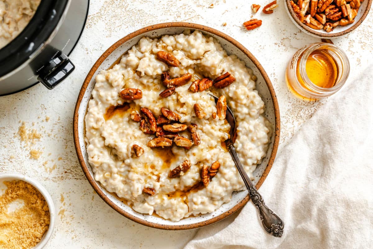 A dish of crock pot pecan pie oatmeal next to a dish of pecans and maple syrup. 