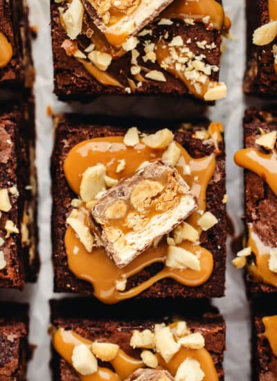 Cut snickers brownies topped with a dollop of caramel, a piece of snickers, and chopped peanuts.