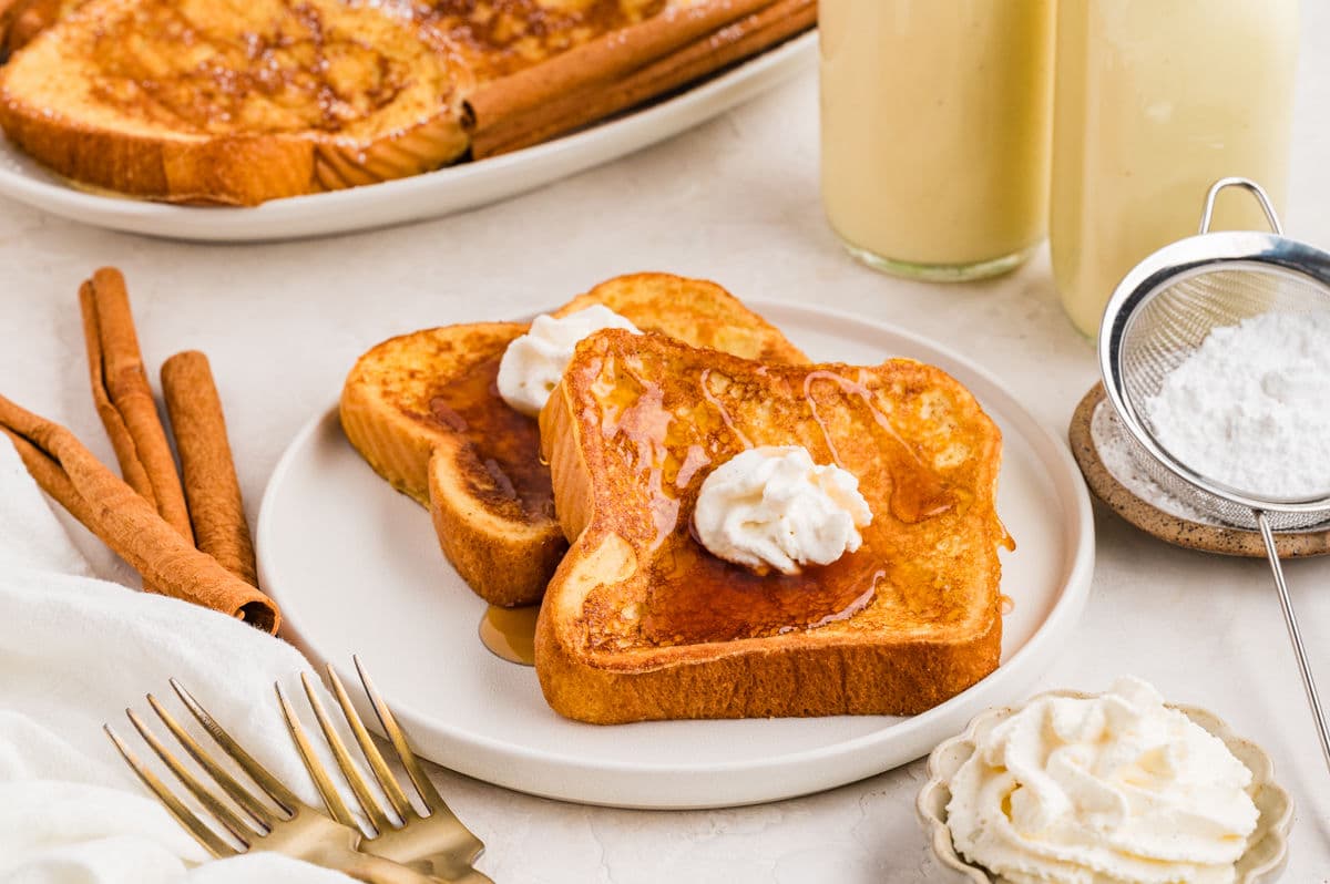 Two pieces of eggnog French toast on a plate topped with whipped cream and syrup.