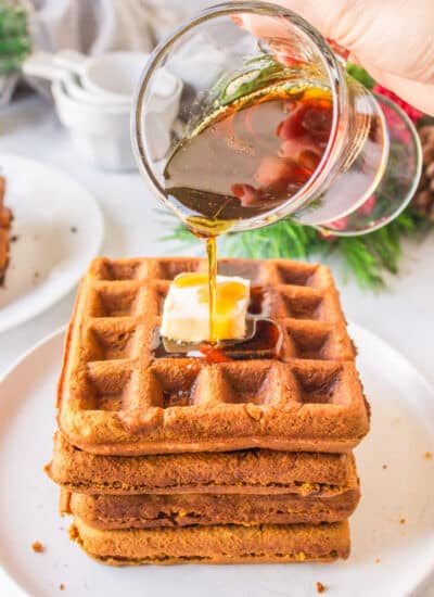 A dish of maple syrup pouring onto a stack of gingerbread waffles.