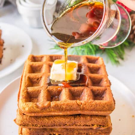 A dish of maple syrup pouring onto a stack of gingerbread waffles.