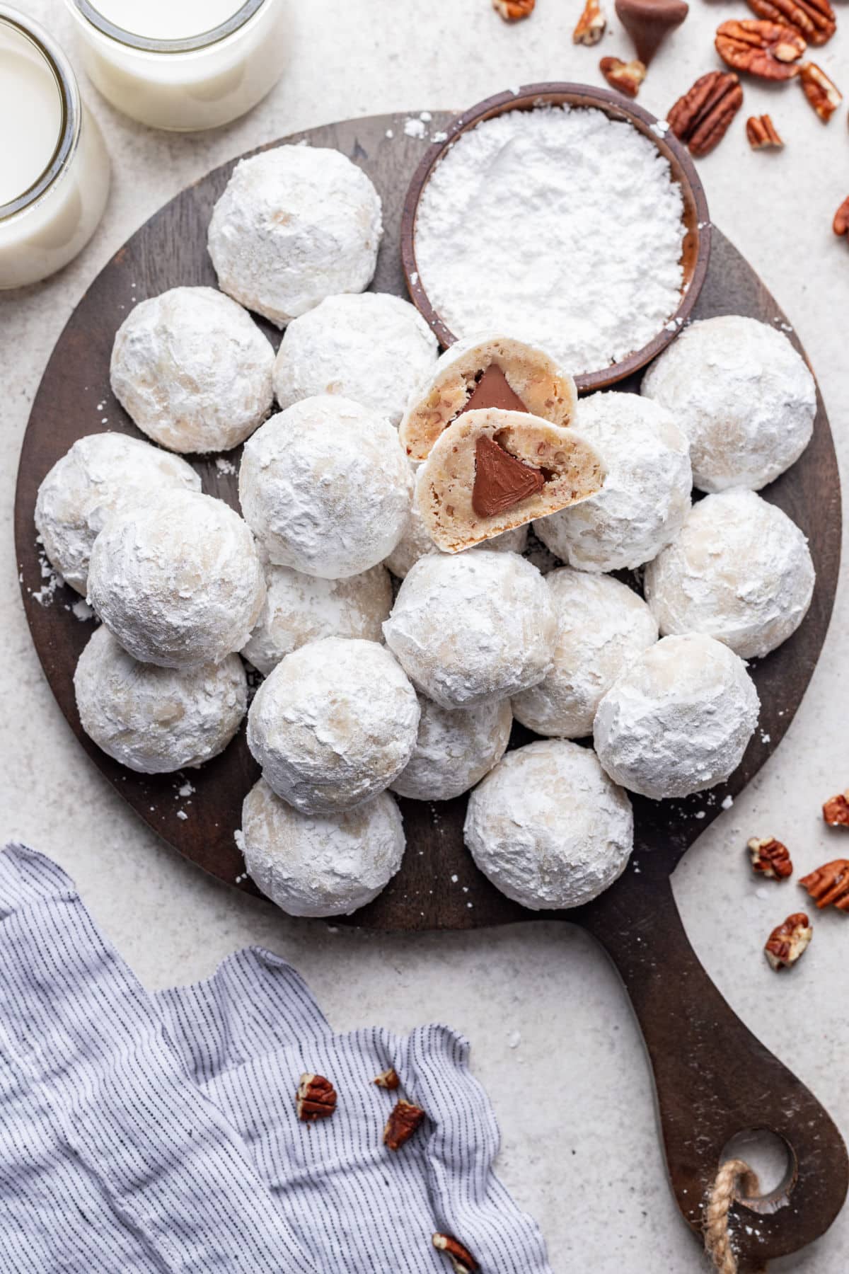 A wooden platter with overlapping stacks of snowball kiss cookies and a dish of powdered sugar.