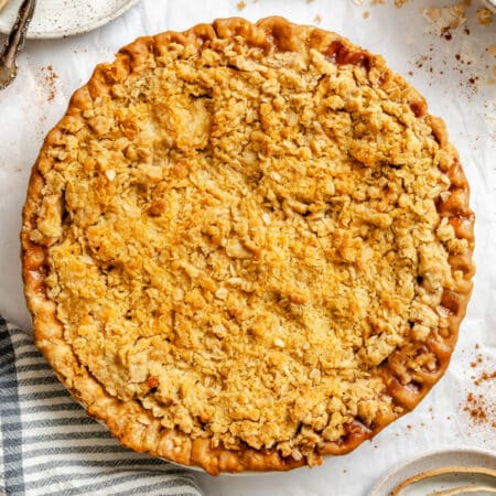 An apple crumble pie next to a stacked plates and silverware.