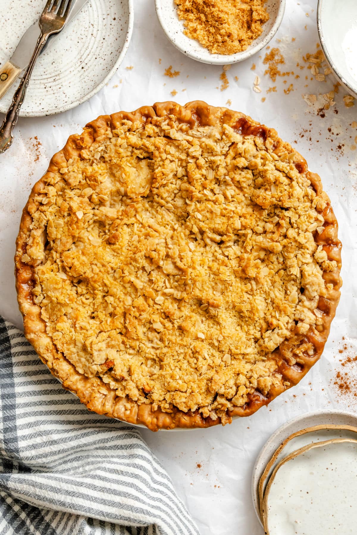 An apple crumble pie next to a stacked plates and silverware.