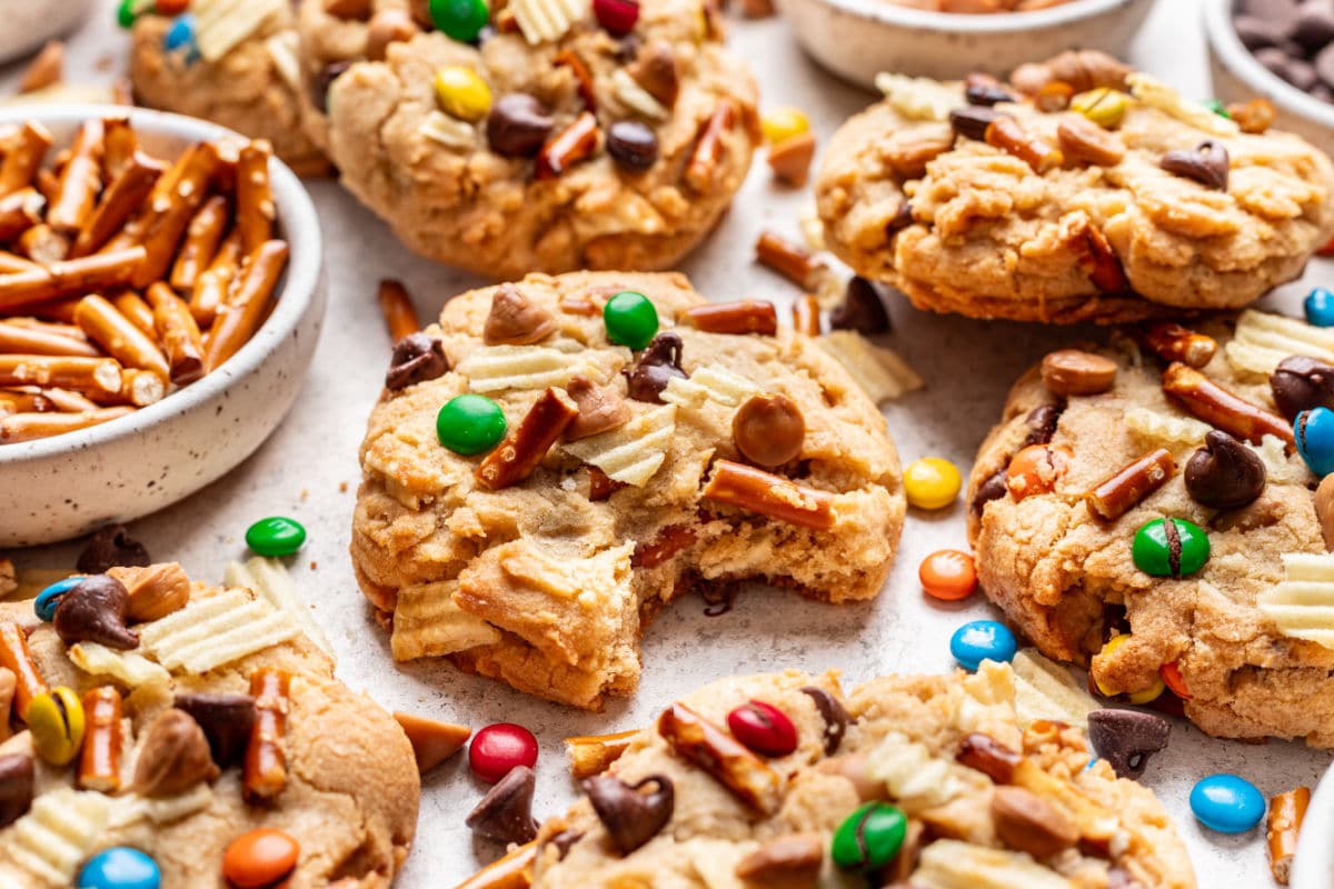 A kitchen sink cookie with a bite missing next to overlapping cookies and a dish of pretzels.