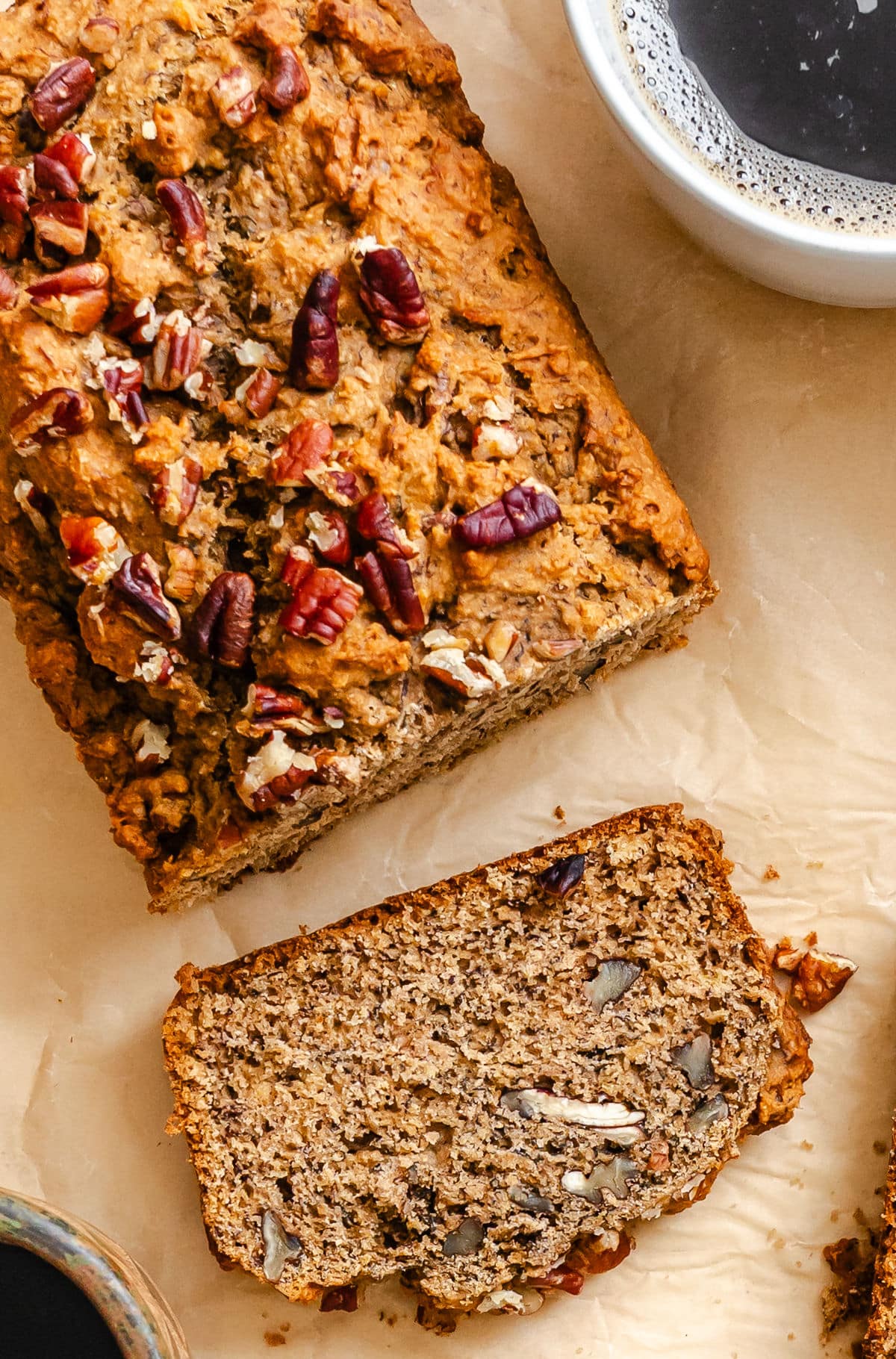 A slice of whole wheat banana bread next to the loaf and a mug of coffee.
