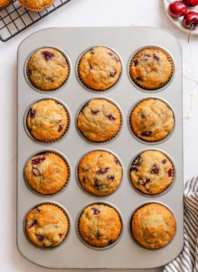 A muffin tin with baked cherry muffins next to a bowl of cherries.