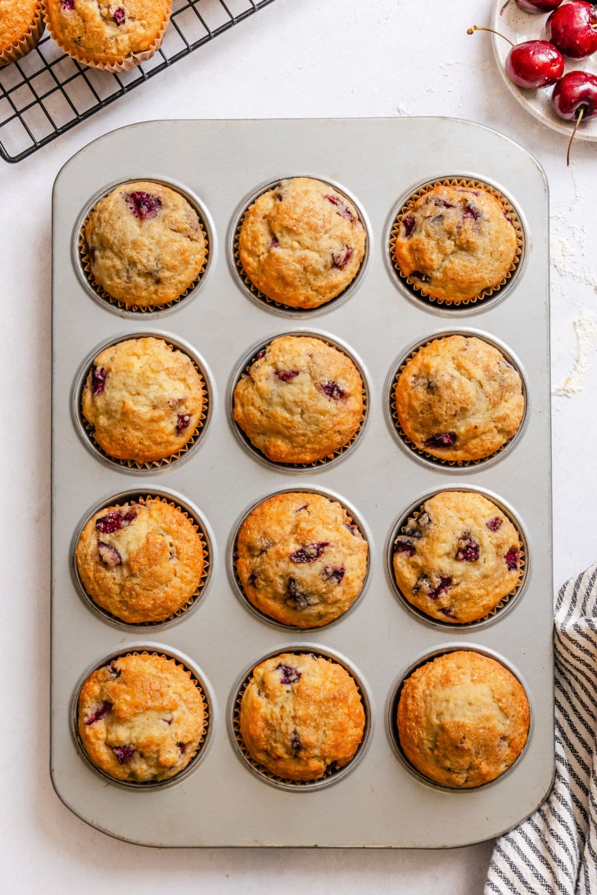 A muffin tin with baked cherry muffins next to a bowl of cherries.