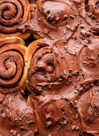 Chocolate frosted cinnamon cinnamon rolls topped with shaved chocolate in a baking pan.