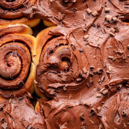 Chocolate frosted cinnamon cinnamon rolls topped with shaved chocolate in a baking pan.