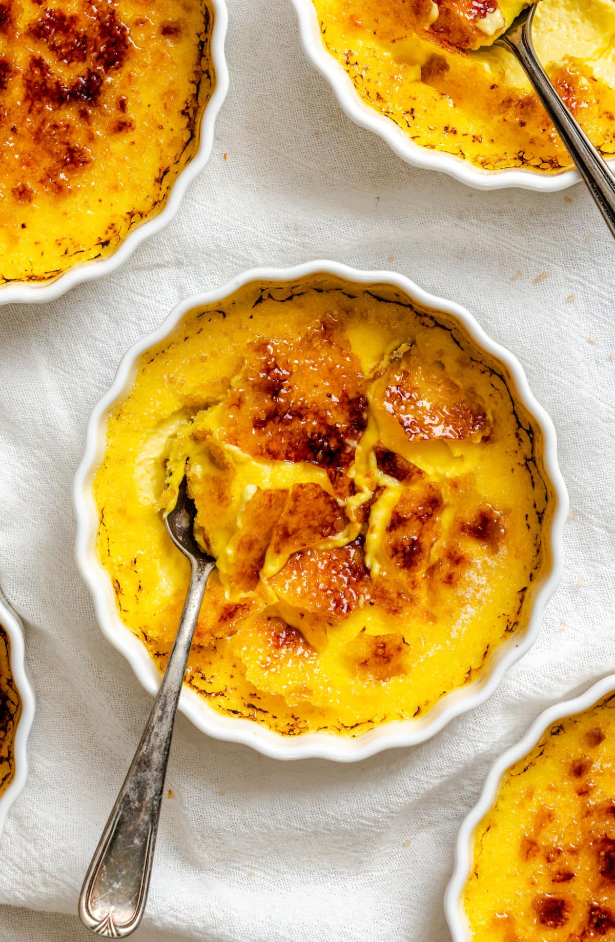 A ramekin of creme brulee with a spoon in it surrounded by more ramekins of creme brulee.