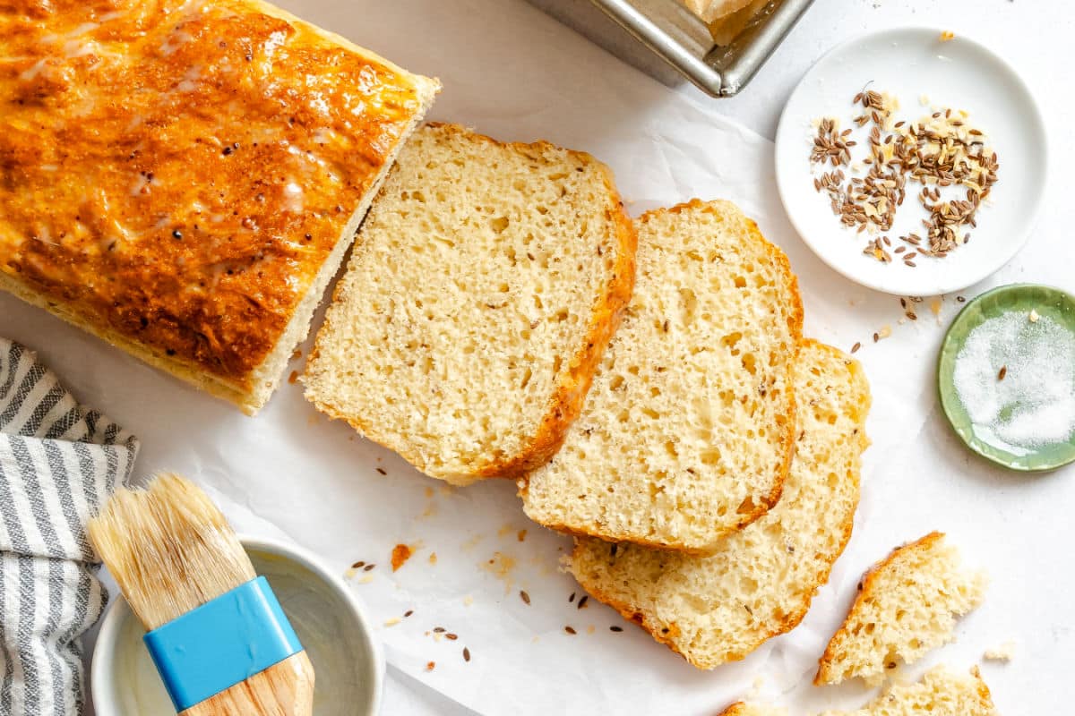 A partially sliced loaf of cottage cheese dilly bread next to a bread pan and a dish of dill seeds. 