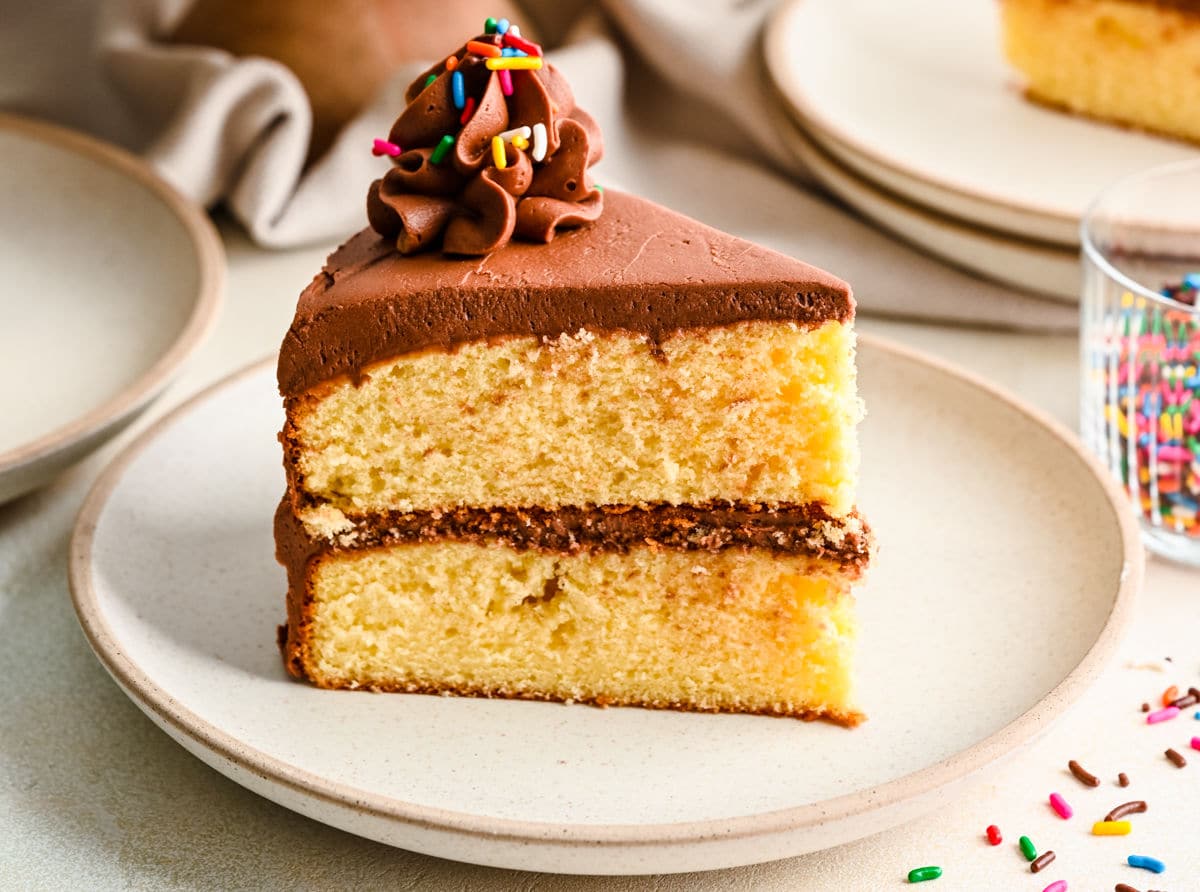 A slice of yellow cake on a cream plate next to a dish of sprinkles. 