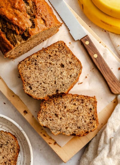 Two slices of banana bread next to the loaf of bread on a cutting board.
