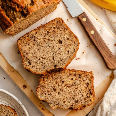Two slices of banana bread next to the loaf of bread on a cutting board.