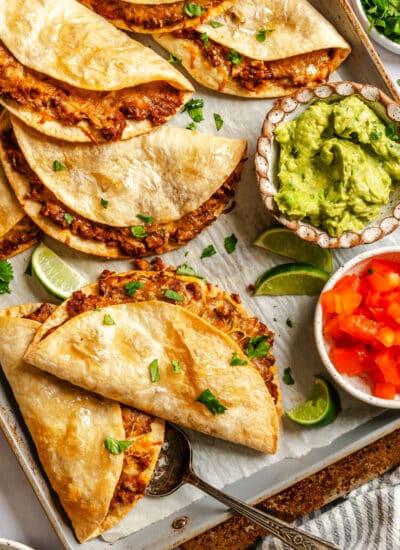 Crispy baked tacos on a baking sheet with dishes of chopped tomatoes and guacamole.