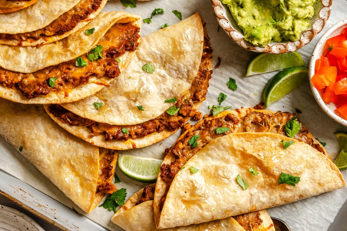baked tacos on a baking sheet next to a piece of lime and a dish of guacamole. 