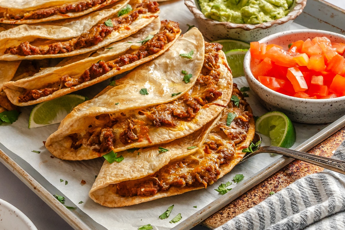 Overlapping baked tacos on a baking sheet next to a striped kitchen towel. 