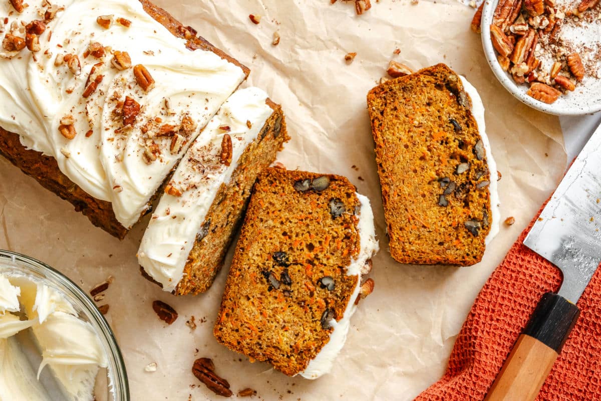 Two slices of carrot loaf cake next to the loaf and a dish of pecans.