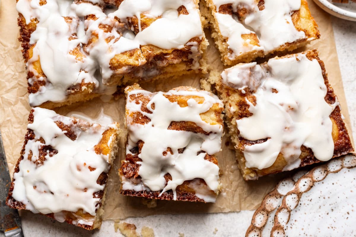 Three slices of cinnamon roll cake next to the cake on brown parchment paper. 